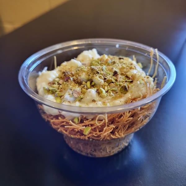 Middle-Eastern dessert in a clear plastic cup, featuring shredded phyllo, cream, and crushed pistachios.